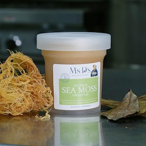 Può includere: Un barattolo di gel cremoso di soursop e muschio marino Ms. D's con coperchio ed etichetta bianchi. L'etichetta include il nome del marchio e la descrizione del prodotto. Accanto al barattolo c'è un mucchio di muschio marino essiccato e alcune foglie secche.