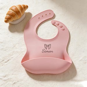 May include: A pink silicone baby bib with a food-catching pocket. The bib has a bow graphic and the name "Simon" printed on it. A golden-brown croissant rests on a white plate in the background.