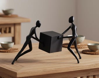 May include: A black metal sculpture depicting two stylized figures pushing a black cube. The figures are elongated and minimalist in design, creating a sense of movement and effort. The sculpture is placed on a wooden table.