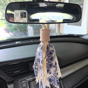 May include: A car rearview mirror hanging ornament featuring a light-colored wooden spool top with a dangling tassel made of multiple strips of fabric. The fabric strips display a blue and white patterned design mixed with plain cream-colored fabric.