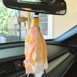 May include: A car rearview mirror hanging ornament featuring a central wooden spool painted yellow with a white floral pattern, adorned with multiple hanging strips of fabric in shades of yellow, peach, cream, and white with frayed edges.