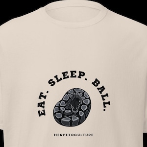 Könnte beinhalten: Beigefarbenes T-Shirt mit einer Grafik einer zusammengerollten Schlange in Grau- und Schwarztönen. Der Text "EAT. SLEEP. BALL." ist über der Schlange angeordnet, darunter steht "HERPETOCULTURE".