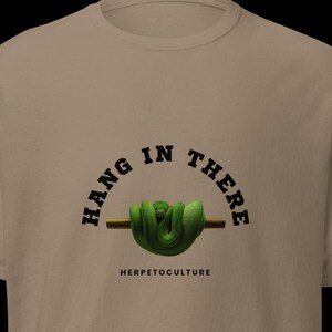 以下が含まれることがあります： ベージュ色のTシャツ。緑色のヘビが茶色の枝に巻き付いたデザイン。ヘビの上には「HANG IN THERE」、下には「HERPETOCULTURE」の文字。半袖、クルーネックのスタイルです。