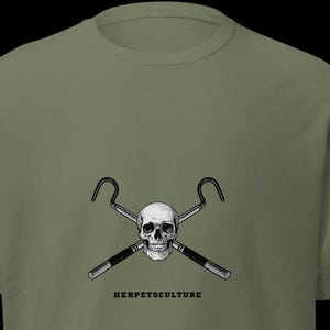 Könnte beinhalten: Olivgrünes T-Shirt mit einem schwarz-weißen Motiv eines Schädels, gekreuzten Schlangenhaken und dem Wort "HERPETOCULTURE". Das T-Shirt hat einen Rundhalsausschnitt und kurze Ärmel.
