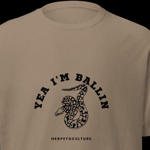 Peut inclure: T-shirt de couleur beige avec un motif noir d'un serpent enroulé, accompagné des mots "Yea I'm Ballin" en arc de cercle et "HERPETOCULTURE" en dessous. Le t-shirt a des manches courtes et un col rond.