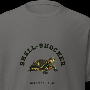 Puede incluir: Una camiseta gris con una ilustración de tortuga y el texto "SHELL-SHOCKER" arqueado encima. La palabra "HERPETOCULTURE" está debajo. La tortuga tiene cabeza y patas verdes y rojas, con un caparazón amarillo y negro.