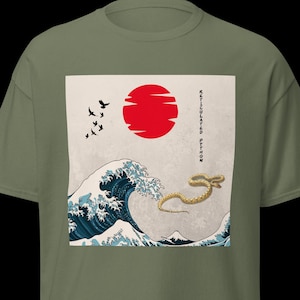 May include: Olive green t-shirt with a graphic print. The design features a red sun, a large ocean wave, and a yellow snake. Black birds and Japanese text are also included. The shirt is made of a soft material.