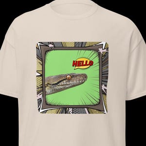 Può includere: T-shirt beige con grafica di una testa di serpente in una cornice in stile fumetto. L'occhio del serpente è visibile e una nuvoletta gialla dice "HELLO". Il disegno è centrato sulla maglietta.