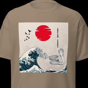 May include: A tan t-shirt featuring a graphic design with a red sun, a blue and white wave, and a stylized cobra. The text "INDIAN COBRA" is printed vertically on the right side of the design. Several black birds are flying in the upper left corner.