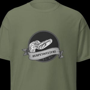 Pode incluir: Camiseta verde-oliva com um design em preto e branco. O design apresenta um crânio de cobra dentro de um círculo preto, com a palavra "HERPETOCULTURE" em uma faixa. A camiseta tem gola redonda e mangas curtas.