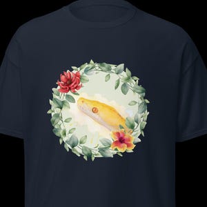 Può includere: T-shirt blu navy con un'illustrazione ad acquerello di un serpente giallo e una ghirlanda floreale. La testa del serpente è centrata all'interno di una ghirlanda di foglie verdi, fiori rossi e arancioni. La maglietta è realizzata in un materiale morbido e confortevole.