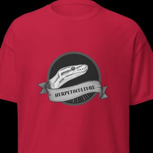 Op de afbeelding: Rood t-shirt met een zwart cirkelvormig embleem. Het embleem toont een gedetailleerde illustratie van een slangenkop en een banner met het woord "HERPETOCULTURE" in een schreefletter. Het t-shirt is gemaakt van een zacht materiaal.