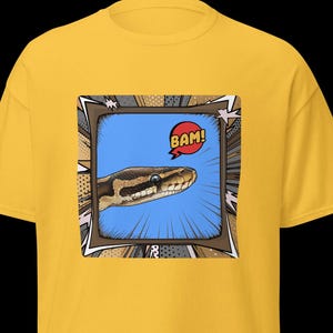 Op de afbeelding: Mosterdgeel T-shirt met een stripboekachtige afbeelding. De afbeelding toont de kop van een slang die uit een blauw kader komt met het woord "BAM!" in een tekstballon. De slang is bruin en beige.