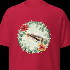 Può includere: T-shirt rossa con un'illustrazione ad acquerello della testa di un serpente circondata da una ghirlanda floreale. Il serpente ha segni marroni e neri. La ghirlanda comprende fiori rossi e gialli e foglie verdi.
