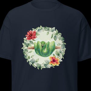 Può includere: T-shirt blu navy con un'illustrazione ad acquerello di un serpente verde avvolto attorno a un ramo, circondato da una ghirlanda floreale con fiori rossi e arancioni. Il disegno è centrato sulla parte anteriore della maglietta.