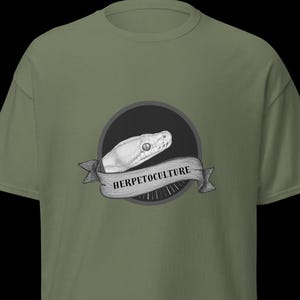 Può includere: T-shirt verde oliva con grafica in bianco e nero di una testa di serpente all'interno di un cerchio e la scritta "HERPETOCULTURE" su uno stendardo. La maglietta è realizzata in materiale morbido.