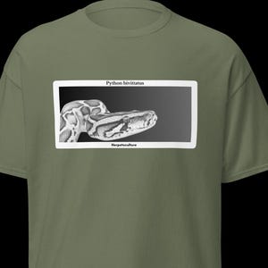Può includere: T-shirt verde oliva con un'immagine in bianco e nero di un serpente. L'immagine è incorniciata in bianco e presenta il testo "Python bivittatus" sopra e "Herpetoculture" sotto. La t-shirt è a maniche corte.