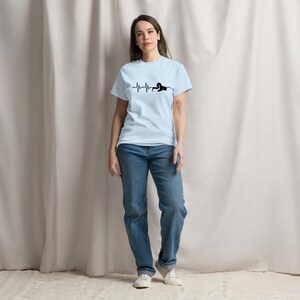 May include: Pale blue t-shirt featuring a black graphic of a heartbeat line and a yoga pose. The model wears blue jeans and white trainers. The background is a neutral-toned curtain. The outfit is casual and comfortable.