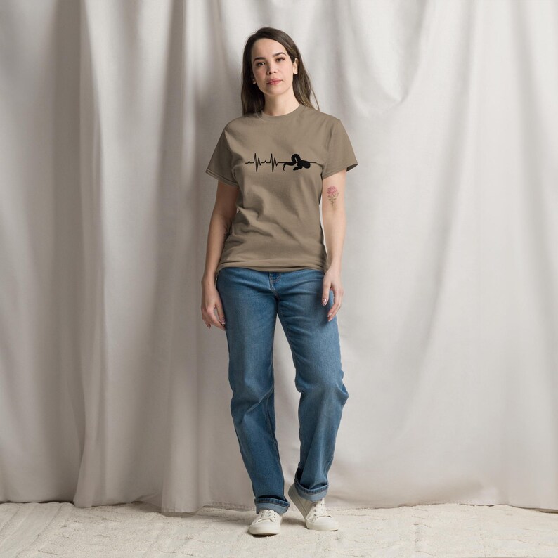 May include: A tan t-shirt with a black heartbeat line design and a person silhouette. The person is wearing blue jeans and white trainers. The background is a white curtain.