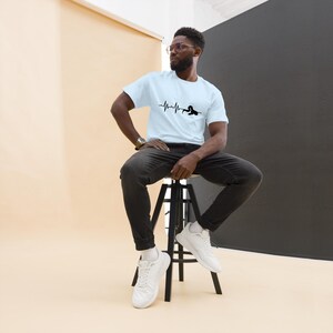 May include: A light blue t-shirt with a black heartbeat line and swimmer graphic. The shirt is made of a soft material. The model is wearing black jeans and white trainers. The image shows a person sitting on a black stool.