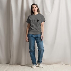 May include: A grey t-shirt featuring a black heartbeat line graphic and a silhouette. The person is wearing blue jeans and white trainers. The t-shirt is a crew neck style.