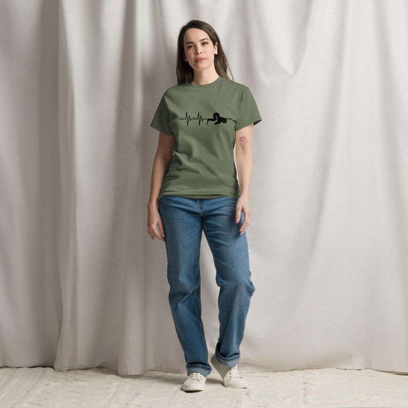 May include: Olive green t-shirt featuring a black heartbeat line graphic and a person silhouette. The person is wearing blue jeans and white trainers. The background is a white curtain.