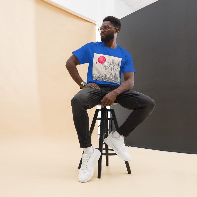 Może przedstawiać: Kr&oacute;lewska niebieska koszulka z grafiką czerwonego słońca nad łodygami bambusa. Koszulka jest noszona przez osobę siedzącą na czarnym stołku. Osoba ta ma na sobie czarne dżinsy i białe trampki.