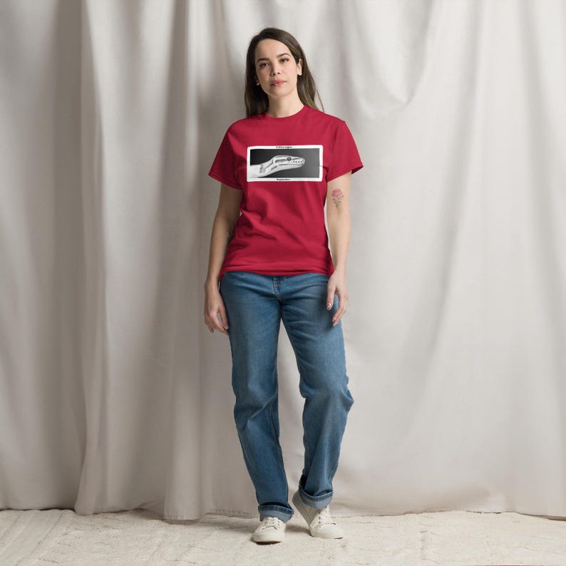 May include: A red t-shirt featuring a black and white snake head graphic within a rectangular border. The person is wearing blue jeans and white trainers.