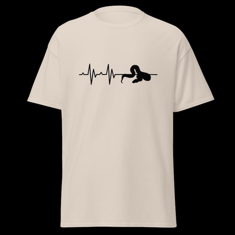 May include: Beige t-shirt with a black graphic depicting a heartbeat line and a snake. The snake is curled up at the end of the line. The shirt has short sleeves and a crew neck. The design is simple and eye-catching.