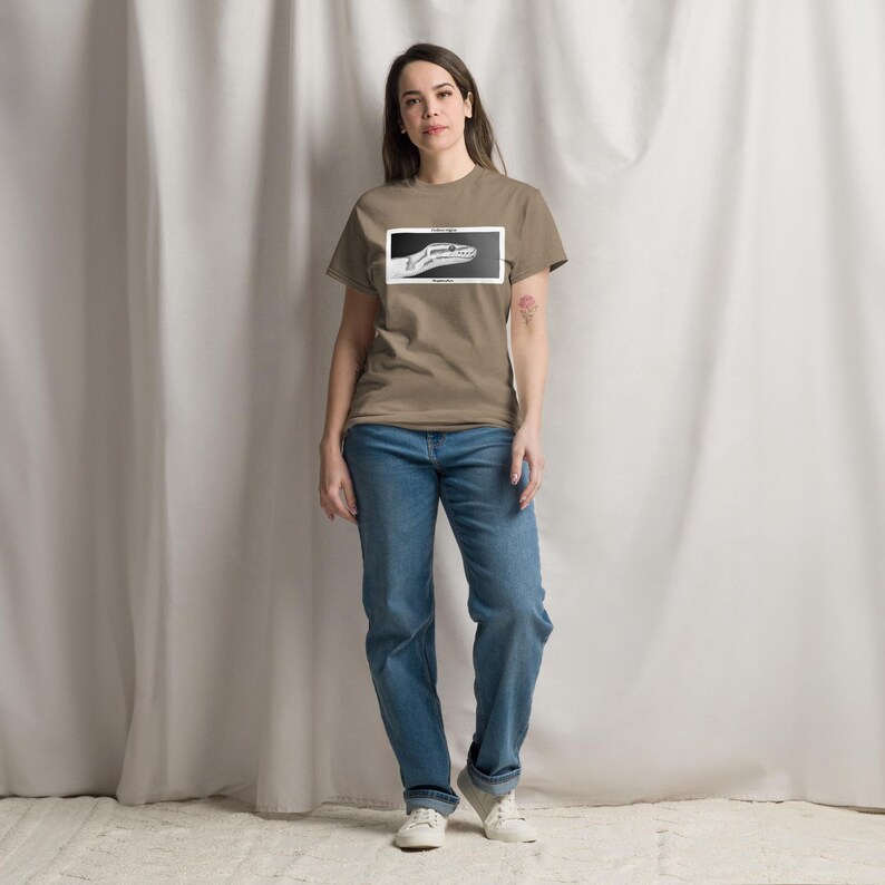 May include: A tan t-shirt with a black and white snake head graphic in a rectangular frame. The model wears blue jeans and white trainers. The shirt design includes text. The image is taken indoors.