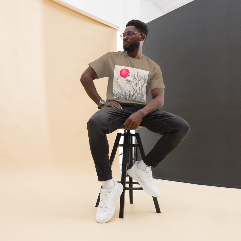 Może przedstawiać: Beżowa koszulka z grafiką czerwonego słońca i łodyg bambusa. Koszulka jest noszona przez osobę siedzącą na czarnym stołku. Osoba ta ma na sobie czarne dżinsy i białe trampki.
