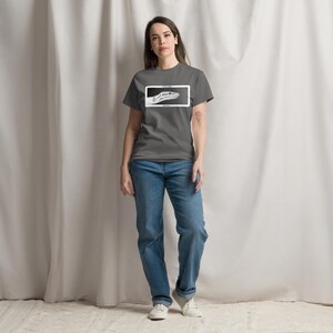 May include: A grey t-shirt featuring a black and white graphic of a snake's head within a white rectangle. The person is wearing blue jeans and white trainers. The shirt is a casual style.