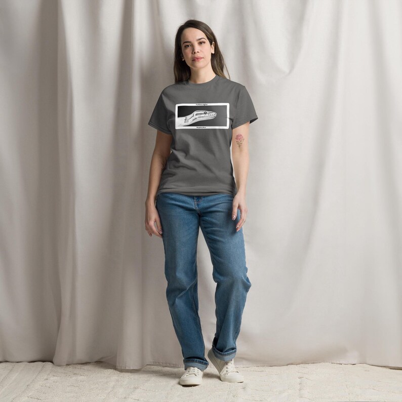 May include: A grey t-shirt featuring a black and white graphic of a snake's head within a white rectangle. The person is wearing blue jeans and white trainers. The shirt is a casual style.