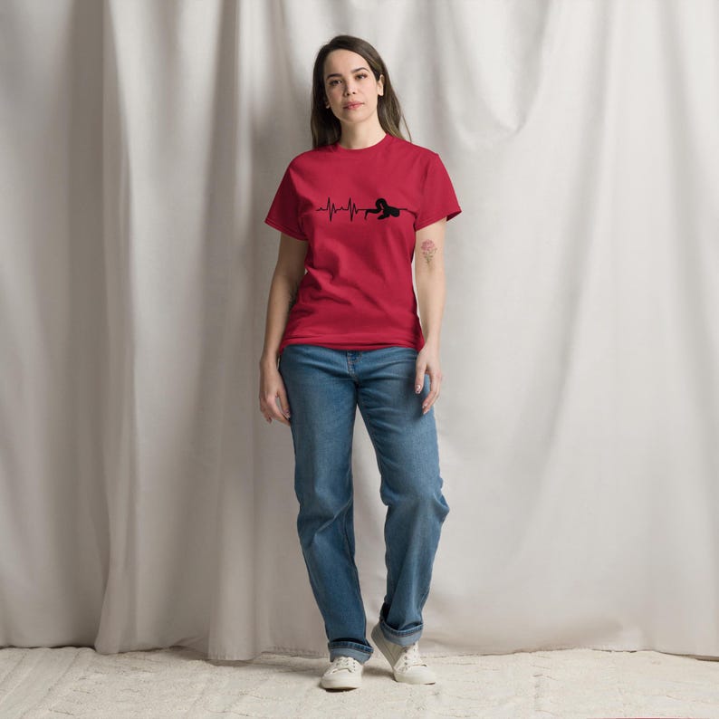May include: A red t-shirt featuring a black heartbeat line design with a silhouette. The person is wearing blue jeans and white trainers. The shirt is made of a soft material.