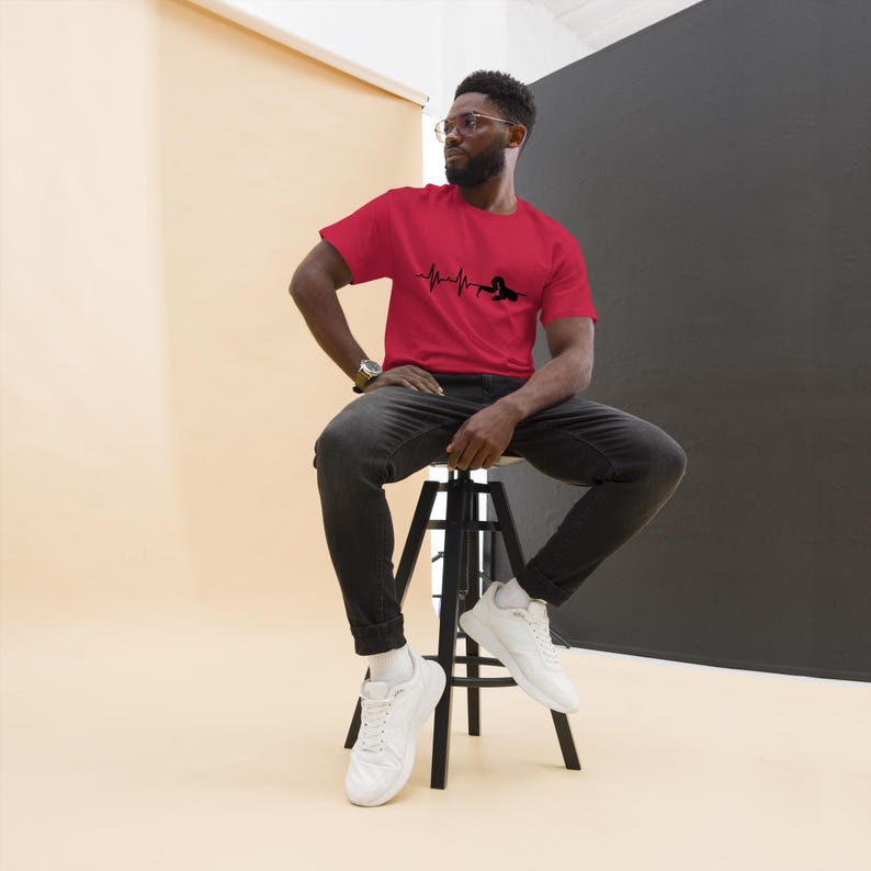 May include: A red t-shirt featuring a black heartbeat graphic and a silhouette. The person is wearing the t-shirt and is sitting on a black stool. The background is a neutral beige and black.