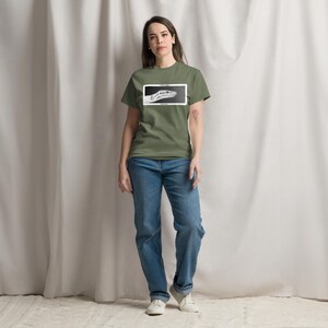 May include: An olive green t-shirt featuring a black and white snake head graphic within a white rectangle. The person is wearing blue jeans and white trainers.