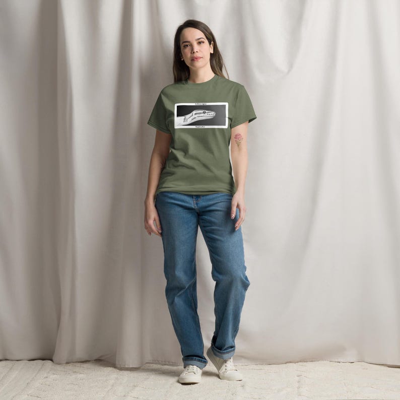 May include: An olive green t-shirt featuring a black and white snake head graphic within a white rectangle. The person is wearing blue jeans and white trainers.