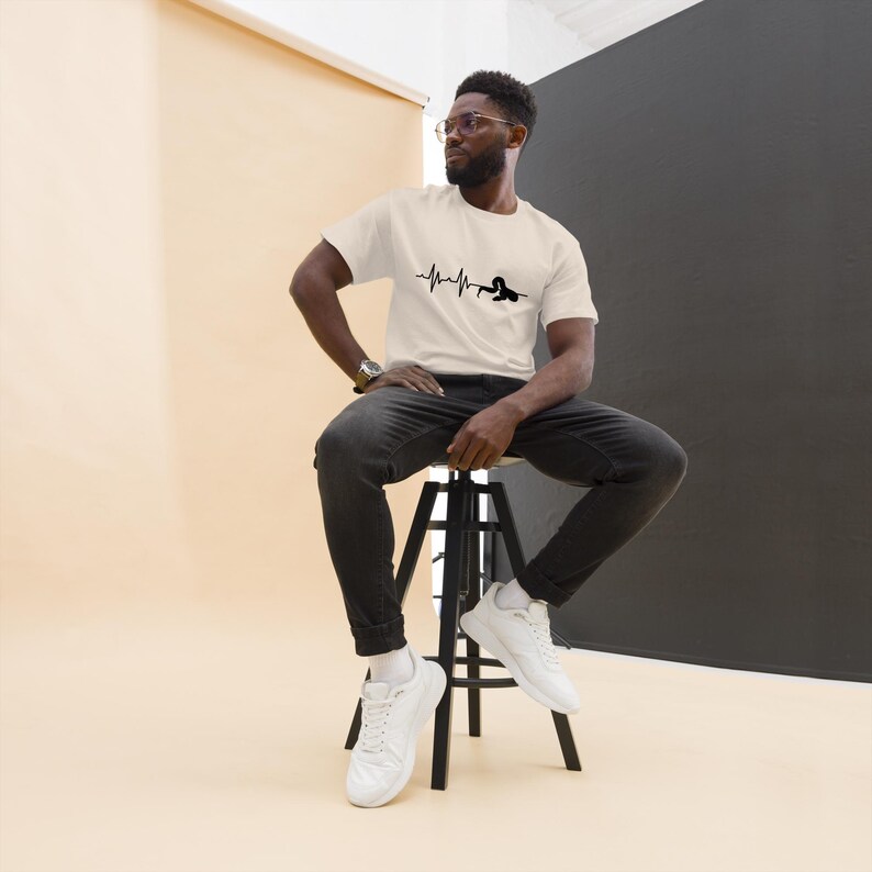 May include: Cream-coloured t-shirt featuring a black graphic of a heartbeat line and a swimmer. The shirt is short-sleeved and worn by a person sitting on a black stool. The person is wearing black jeans and white trainers.