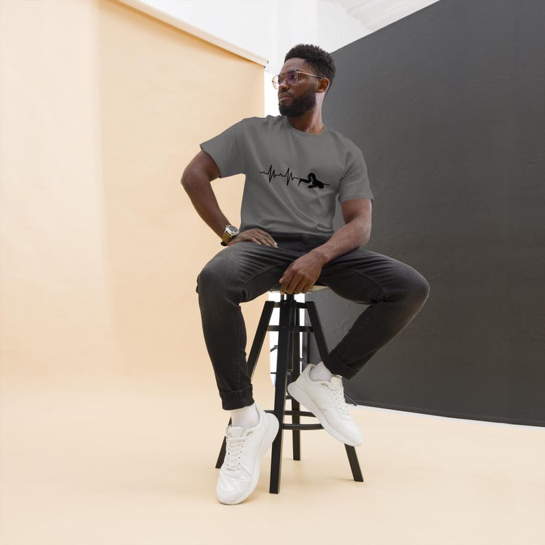 May include: Grey t-shirt featuring a black heartbeat design. The short-sleeved shirt is made of a soft material. The person is wearing black jeans and white trainers.