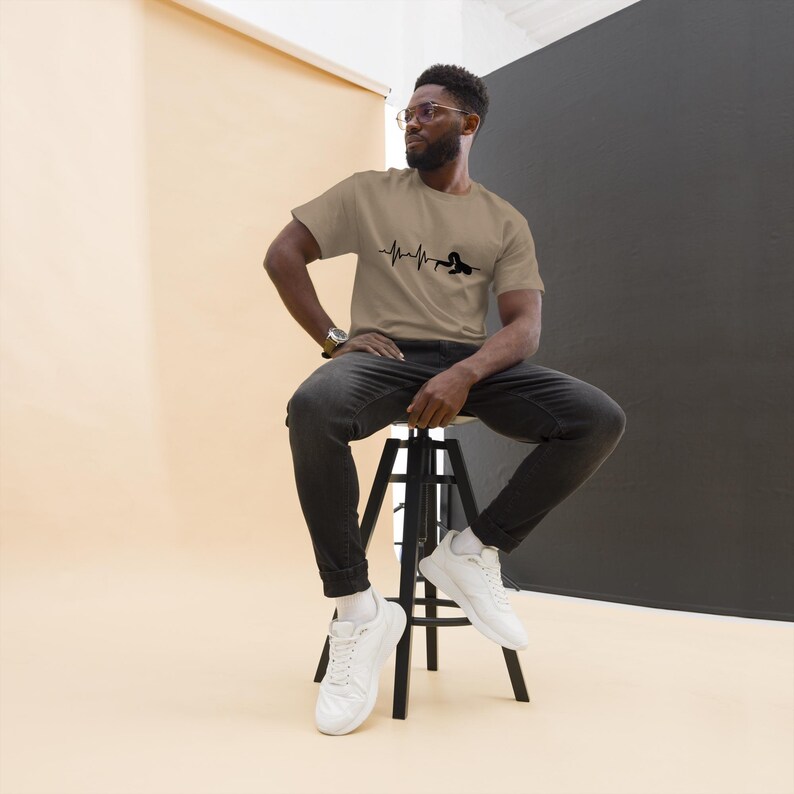 May include: A tan t-shirt with a black heartbeat line design and a silhouette. The shirt is made of a soft material and is being worn by a person sitting on a black stool. The person is wearing black jeans and white trainers.