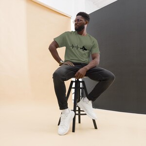 May include: An olive green t-shirt with a black heartbeat graphic and a swimmer silhouette. The person is wearing black jeans and white trainers. The person is sitting on a black stool.