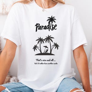 Peut inclure: Un t-shirt blanc à manches courtes avec un motif noir représentant des palmiers, une chaise de plage et un parasol. Le texte Paradise est inscrit au-dessus, et en dessous, on peut lire, That's nice and all... but i'd rather have another snake.