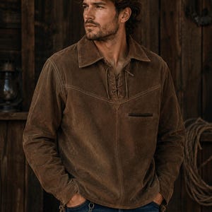 May include: A brown suede pullover shirt featuring a collared neckline with a lace-up front closure, long sleeves, and a single horizontal chest pocket. The garment is paired with blue denim jeans that include a metal chain accessory at the waist.