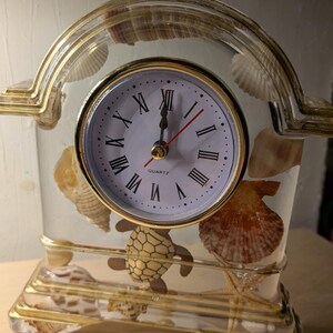 May include: A clear resin mantel clock with a gold-toned frame, featuring a white face with black Roman numerals and the word QUARTZ. The interior is embedded with various seashells, a starfish, and a small turtle figure, creating a coastal theme.