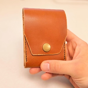 May include: A small, rectangular, brown leather case with a flap closure and a brass button. The case is held in a hand, showcasing the fine stitching and the natural texture of the leather. The design is simple and classic.