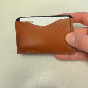 May include: A handmade brown leather card holder with a black interior. The card holder is stitched with black thread and holds a white card. The card holder is held by a hand.