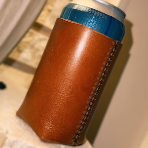 May include: A brown leather can cooler with a stitched seam detail. The cooler holds a blue and silver can. The leather has a glossy finish and a natural color.