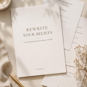 May include: A stack of off-white paper with the text "REWRITE YOUR BELIEFS" and "A short guided exercise to shift your mindset". Additional sheets with prompts and the text "CHALLENGE THAT BELIEF" are visible. A gold pen and dried flowers are also present.