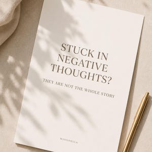 May include: A rectangular white paper card with dark gray text that reads STUCK IN NEGATIVE THOUGHTS? THEY ARE NOT THE WHOLE STORY and MAISONELLA at the bottom. A gold-colored metal pen is positioned to the right of the card on a light surface.