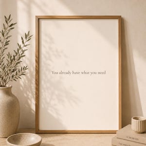 Puede incluir: Una lámina de arte mural rectangular en un marco de madera clara con el texto You already have what you need en una fuente oscura sobre un fondo crema. La lámina se muestra junto a un jarrón de cerámica con ramas de olivo y un cuenco.